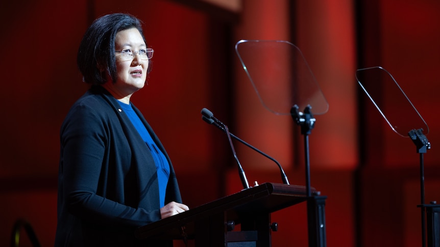 Awoman standing at a podium delivering a speech. She is wearing glasses and a dark outfit with a blue top underneath. 