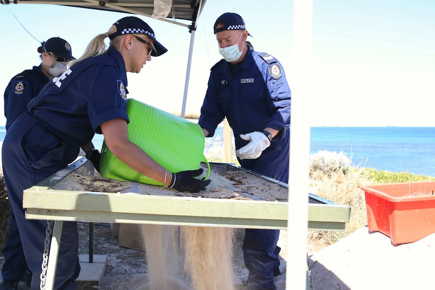 Three forensic officers standing under a temporary cover, sifting sand through a grate.