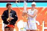 Iga Swiatek holds up a trophy while Naomi Osaka smiles in the background.