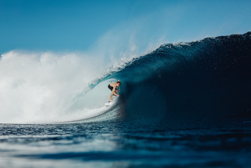 Brett Connellan surfs a large barreling wave, shirtless and wearing black board shorts.