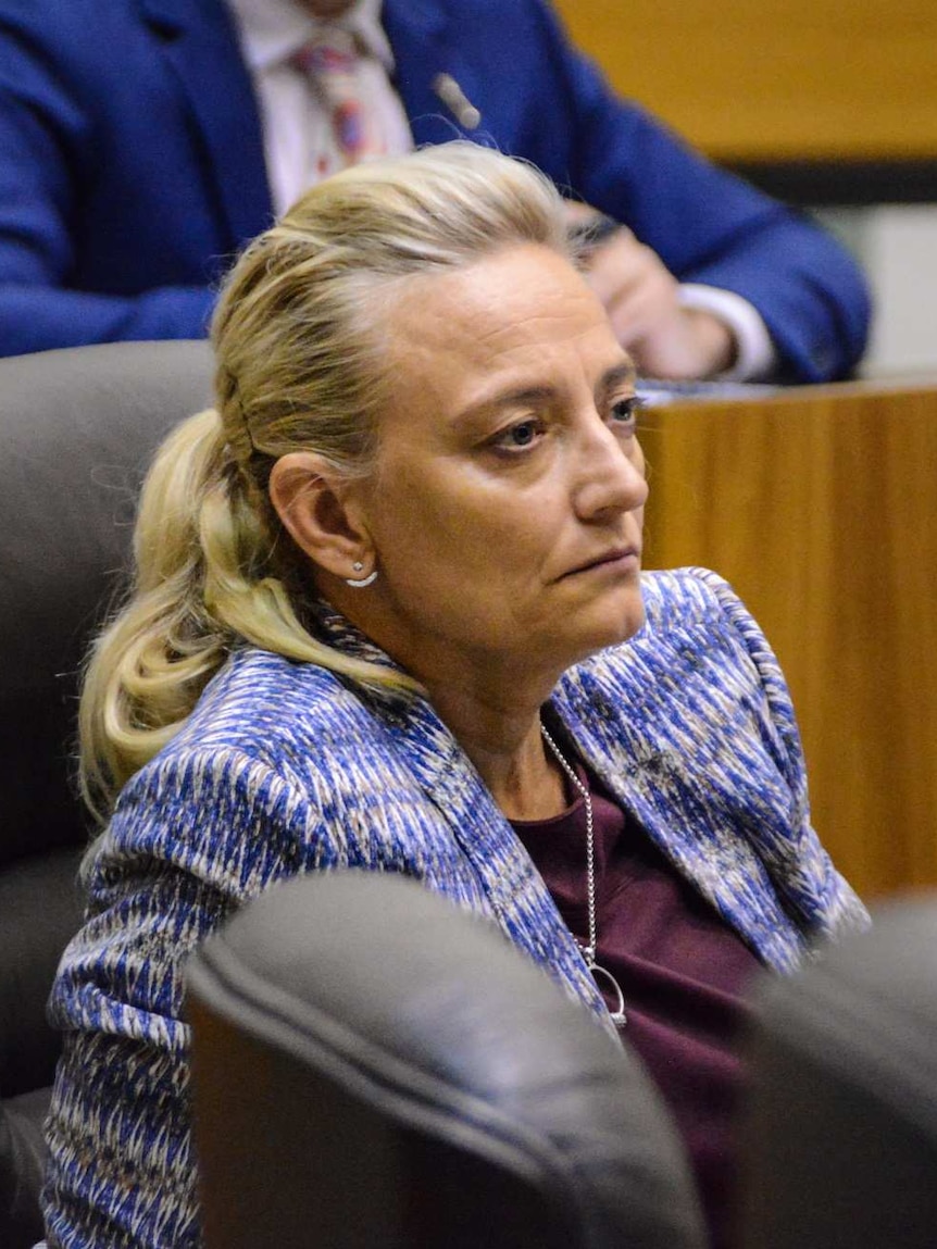 A blonde woman sits in a parliament building with a serous look on her face.