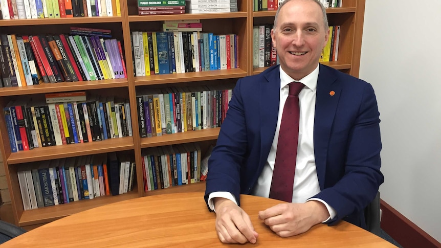 A smiling middle-aged man in a suit and tie sits an office full of books