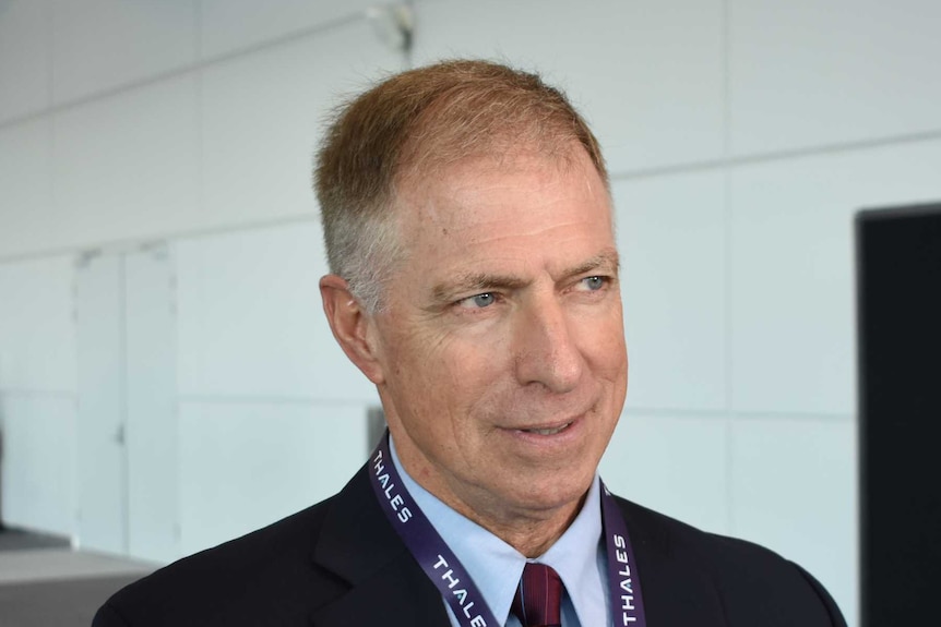 Grant Newsham wears a suit with a maroon tie. He is talking and his hand is slightly raised.