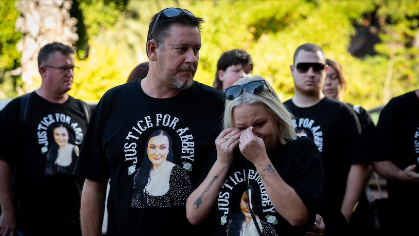 Two people stand in front of a group of people wearing black memorial t-shirts while crying