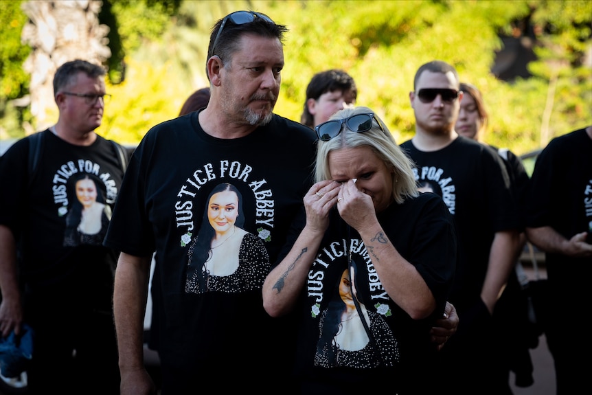 Two people stand in front of a group of people wearing black memorial t-shirts while crying