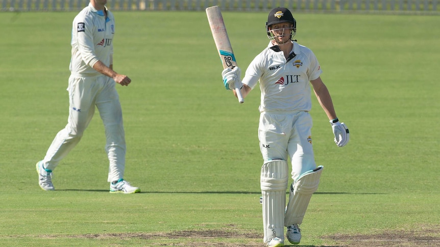 A cricketer raises his bat after making 50 in a Sheffield Shield game, as a fielder walks behind him.