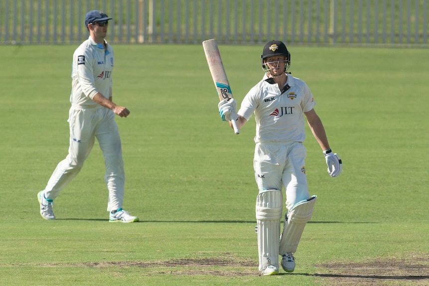 A cricketer raises his bat after making 50 in a Sheffield Shield game, as a fielder walks behind him.