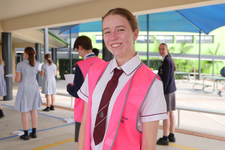 A high school girl stands with a choir outside, wearing a pink COVID-19 safety vest over a white school shirt.