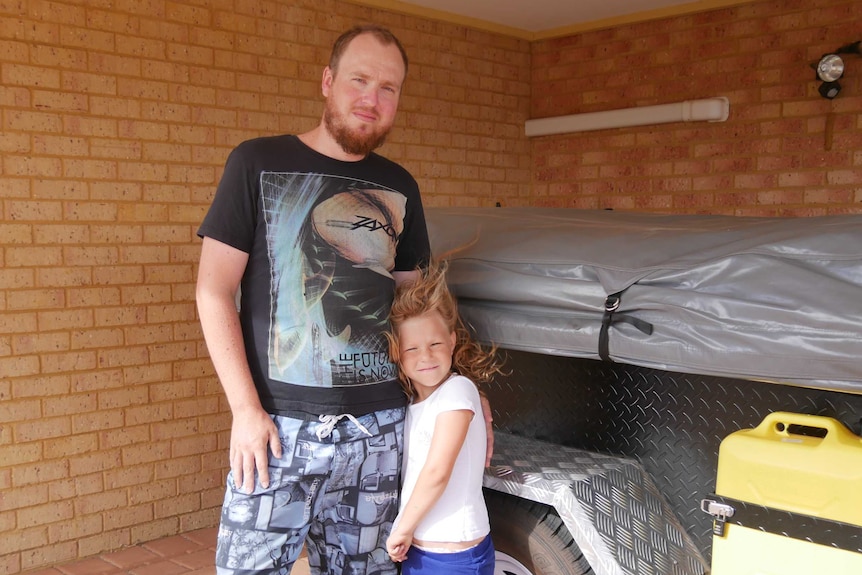 A man in board shorts and a young girl stand next to a trailer in a carport.