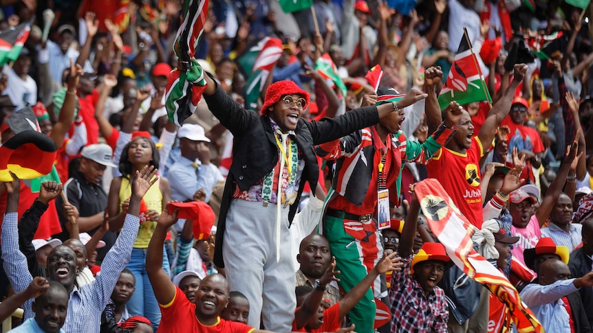 A man stands up in a crowded stadium and looks excited as he raises both arms in the air.