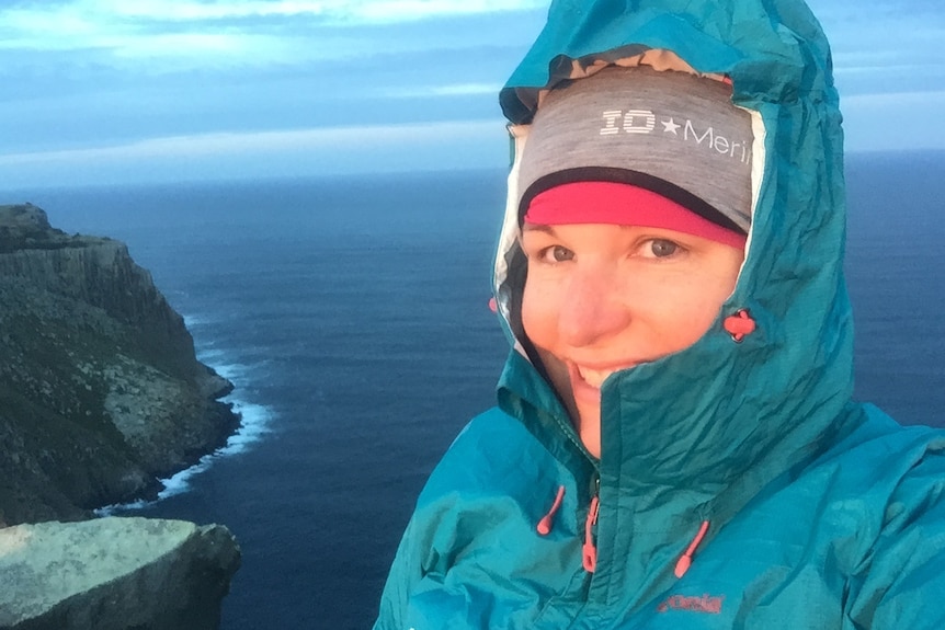 Richelle Olsen wears a beanie and wet weather jacket and standing in front of the ocean, with a cliff behind her.