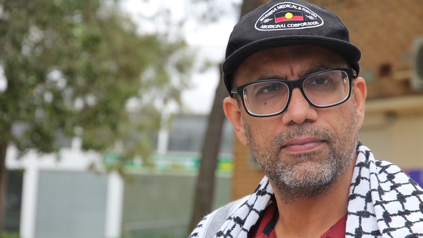 A man with a cap, glasses and wearing a keffiyeh stares past the camera