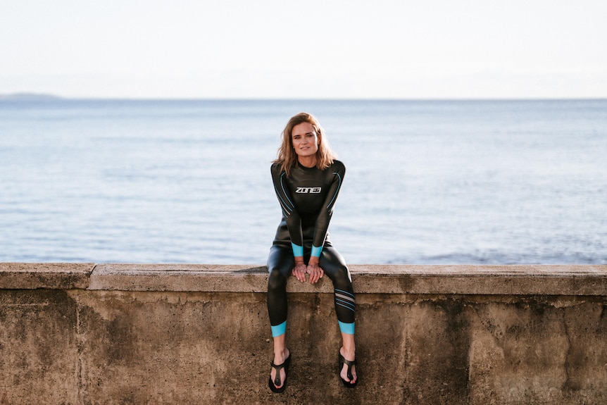 Julia Baird, who has short brown-blonde hair and is wearing a black wetsuit, sits on a sea wall, a vast blue ocean behind her