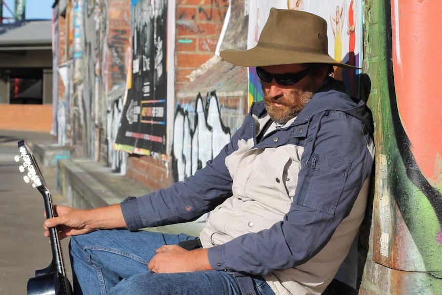 man slumped against graffiti wall, with sunglasses on, and guitar