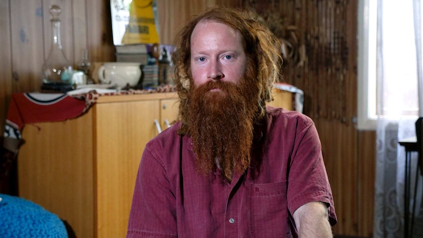 A man in a red shirt with a long beard poses for a photo during an interview in his lounge room.