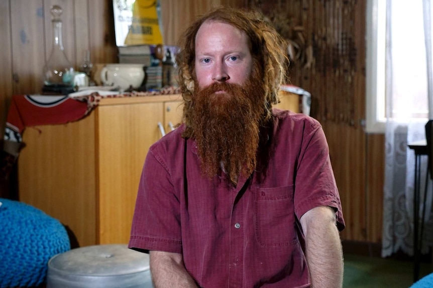 A man in a red shirt with a long beard poses for a photo during an interview in his lounge room.