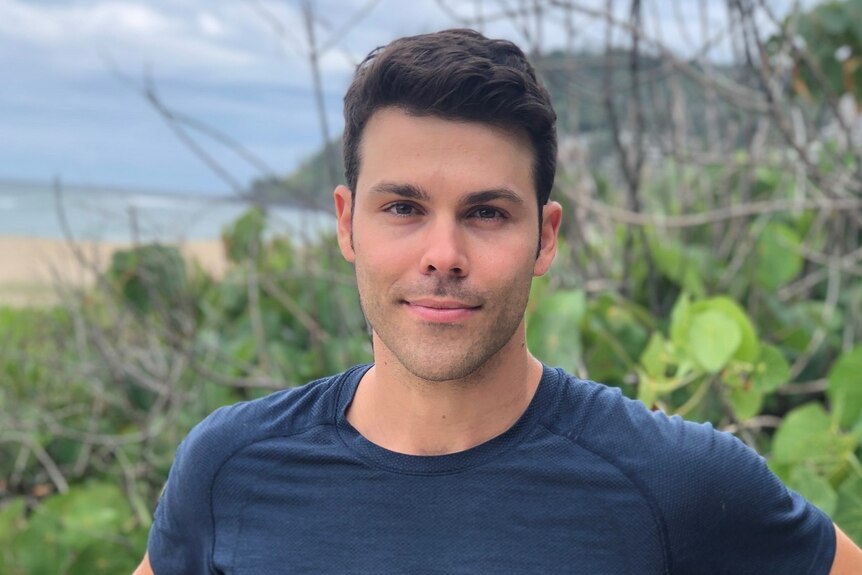 A portrait photo of Aaron Smyth leaning backwards against a wooden fence with green foliage and a beach in the background.