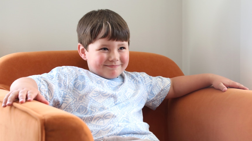 A boy with a cheeky grin on his face sits on an orange armchair.