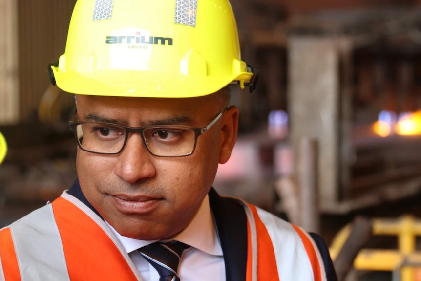 A bespectacled man wearing a hard hat and a high-vis vest over a shirt and tie standing in an industrial area.