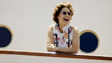 HM The Queen leans over the banister of Royal Yacht Britannia in a floral collared singlet smiling with sunglasses on.
