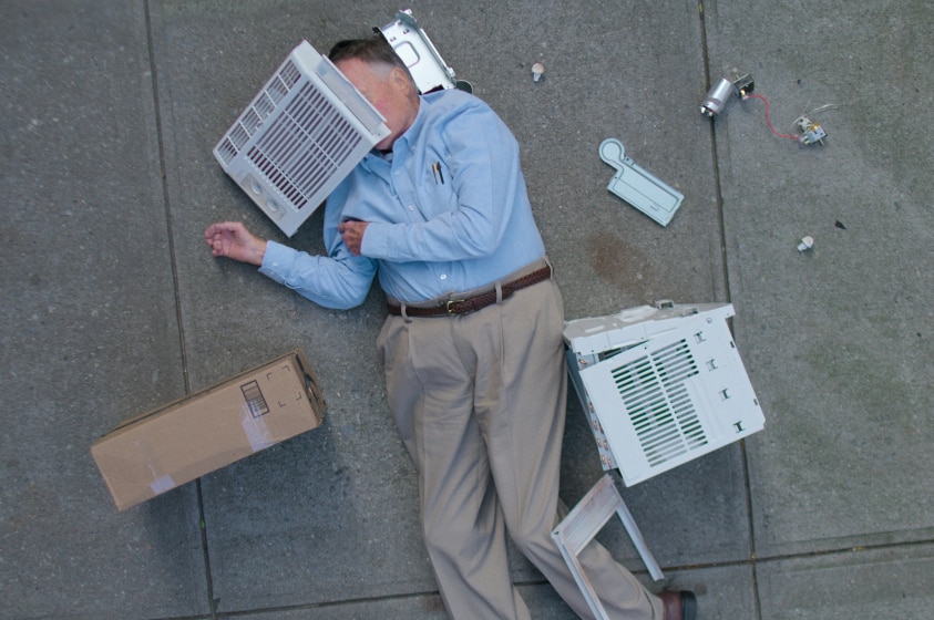An older man in khaki slacks and blue shirt lies on back in unnatural pose on pavement surrounded by air conditioner parts.