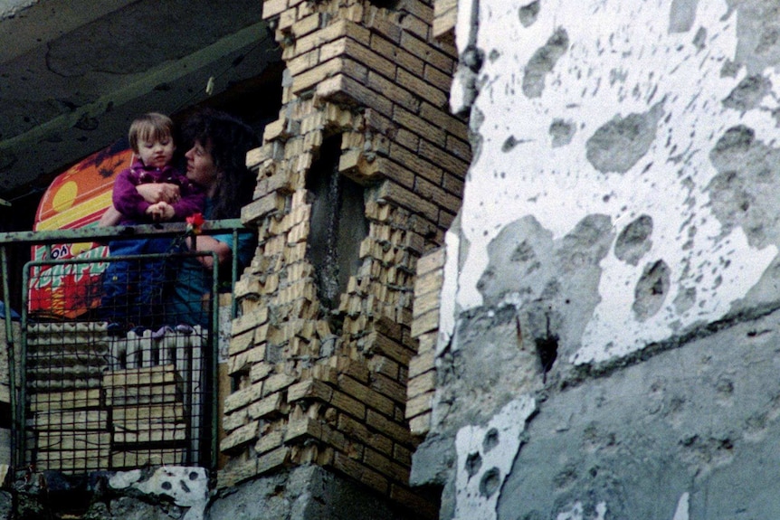 A woman and young child stand on the balcony of a badly damaged building riddled with bullet holes.