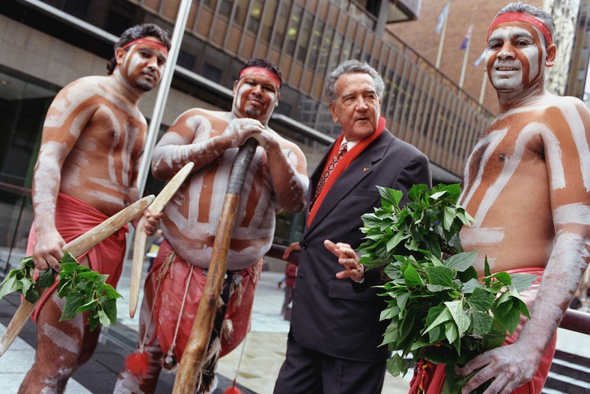 Charles Perkins dressed in a grey suit talking to three Indigenous men holding leaves, boomerangs and a digeridoo.
