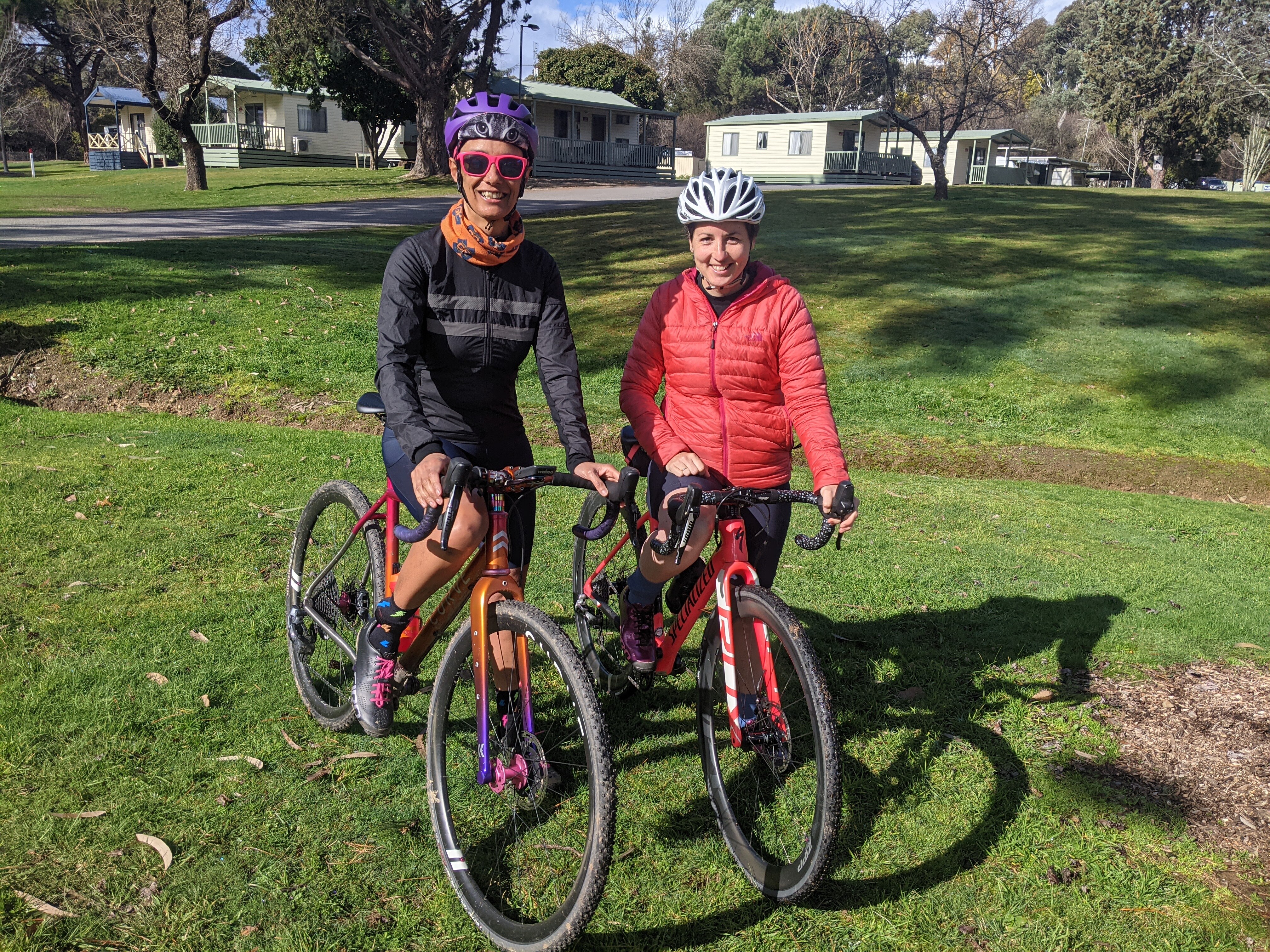 Two women wearing helmets and bright clothes sit on their bikes, smiling. 