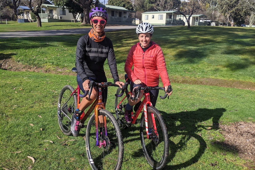 Two women wearing helmets and bright clothes sit on their bikes, smiling. 