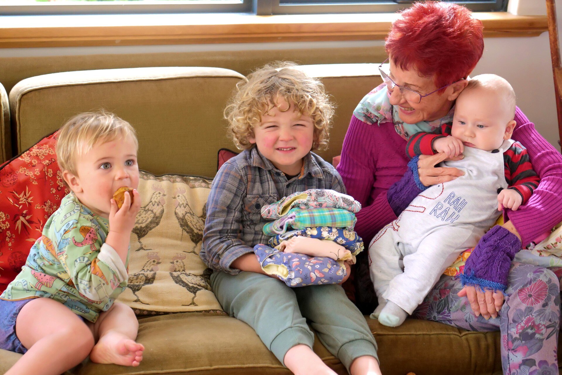 Two toddlers sit in the sunshine on a sofa next to a woman holding a baby.