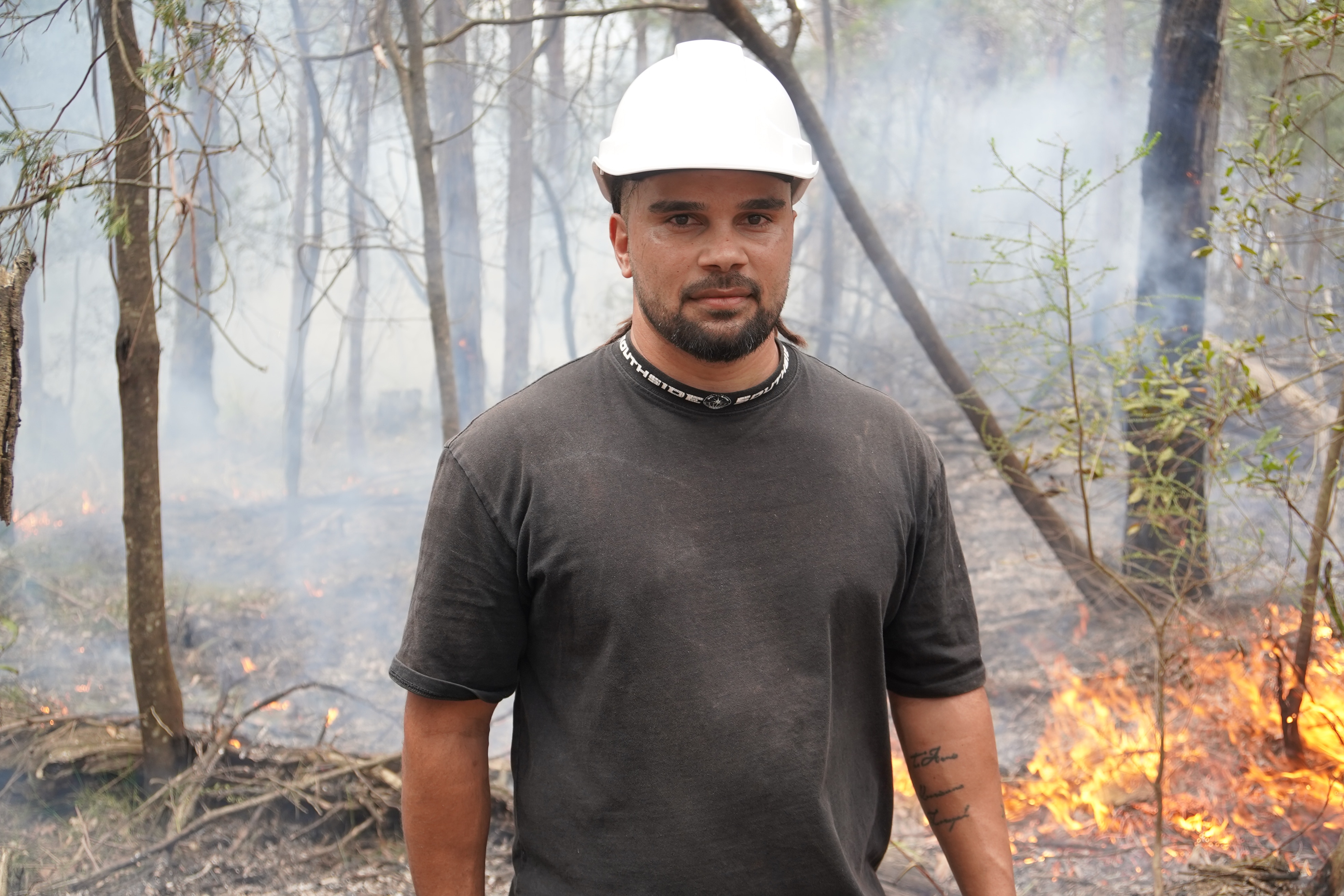 A man with a hard hat looks into the camera with controlled fire behind him.