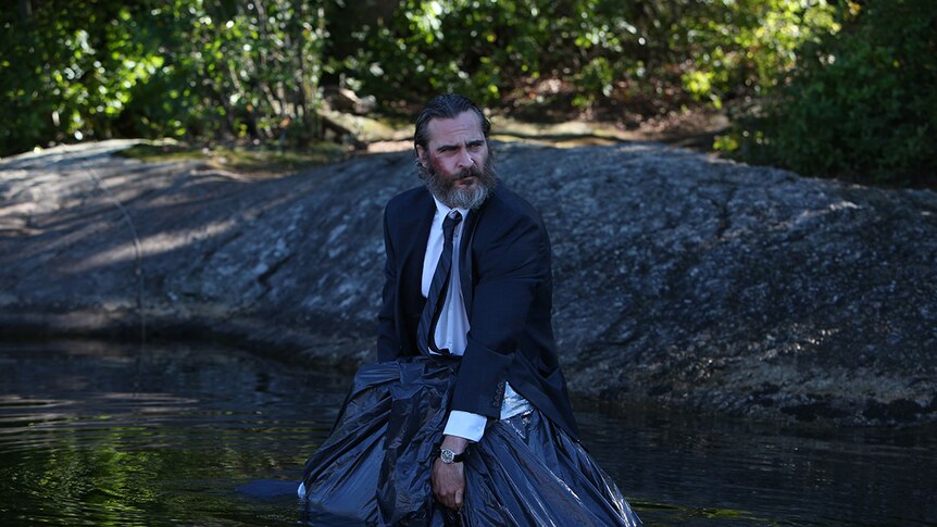 Joaquin Phoenix standing in a river wearing a suit and holding a body bag in film You Were Never Really Here.