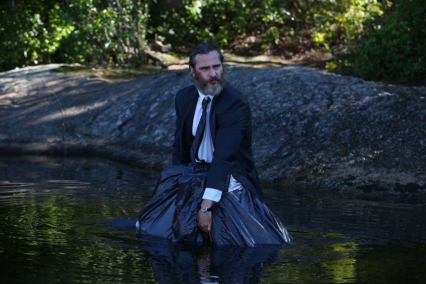 Joaquin Phoenix standing in a river wearing a suit and holding a body bag in film You Were Never Really Here.