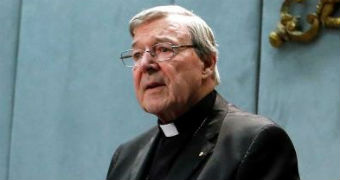George Pell stands before a microphone, hands crossed over each other.