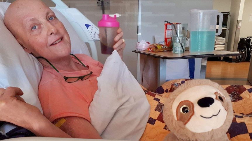 A woman with no hair after chemotherapy lays in a hospital bed, holding a drink and just managing a smile.