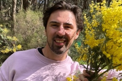 Naarm/Melbourne psychologist Chris Cheers wears a pink jumper and poses beside some flowering wattle in the bush.