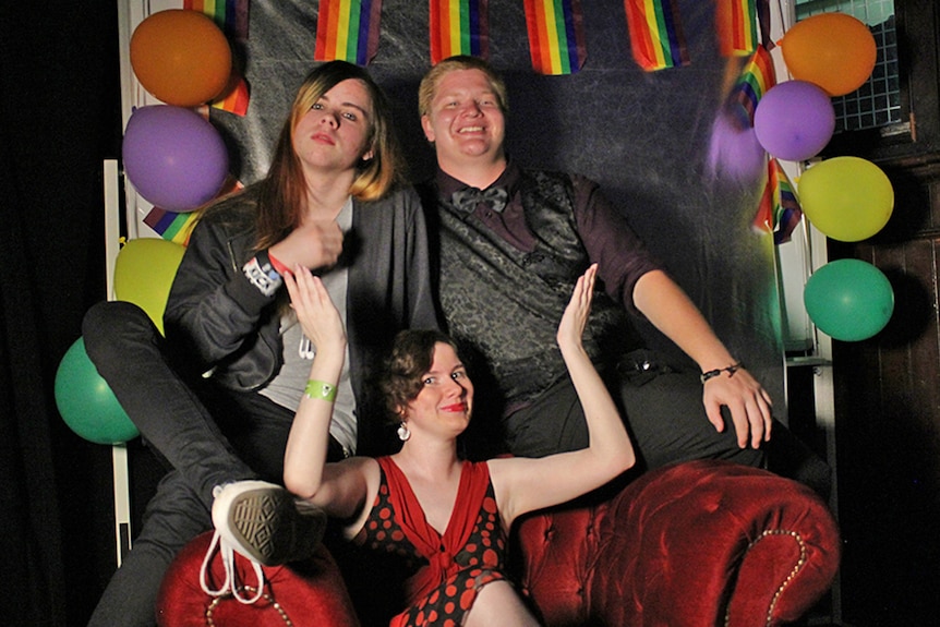 A young woman sits in a velvet chair with two young men seated behind her, rainbow flags and balloons are in the background.