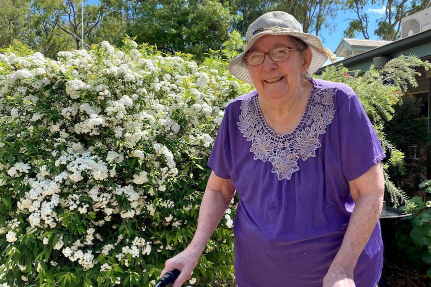 An elderly lady wearing a purple shirt and a hat stands in a garden smiling, using a walker for support.
