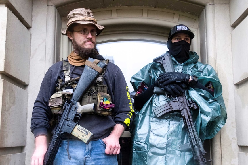 Protesters with long guns shelter from heavy rain by standing in a doorway
