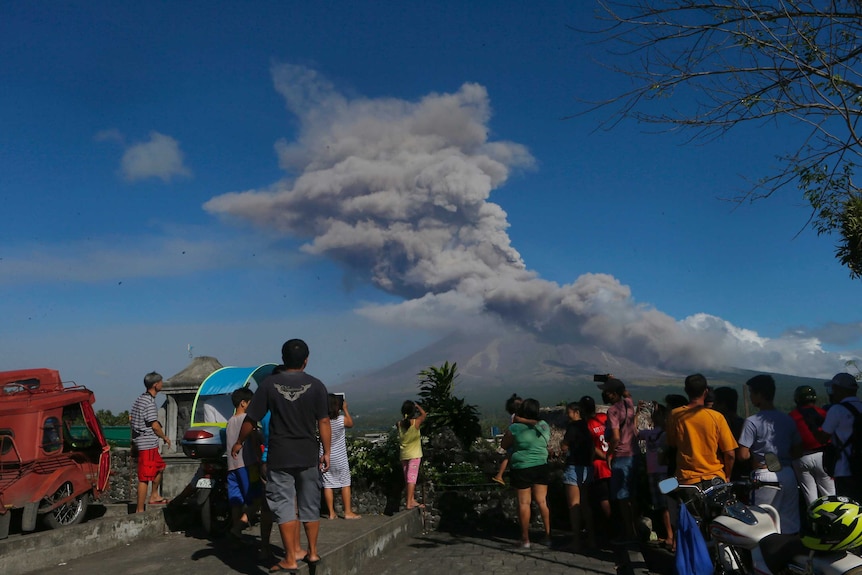 A crowd of people stands at watches the ash cloud from the Mayon volcano in the distance