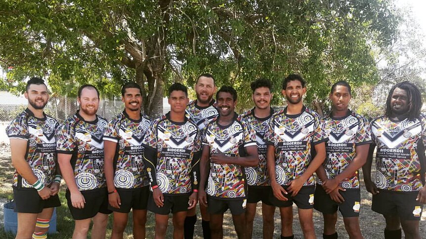Ten Indigenous rugby union players stand in uniform in a row, under a tree