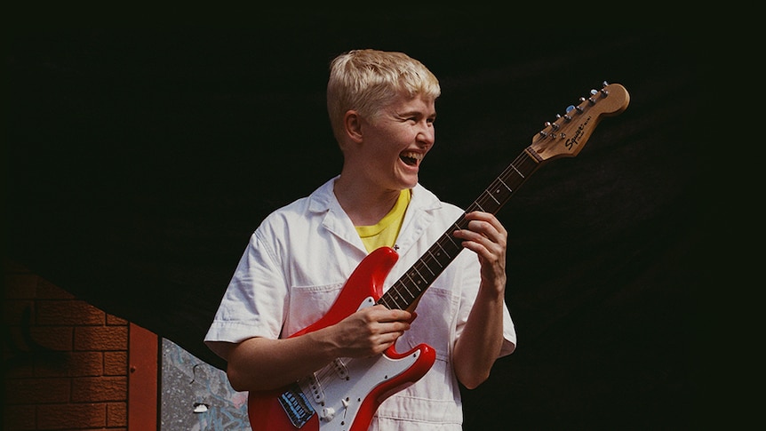 Alex the Astronaut, with short blonde hair and a white button-up, holds a red guitar and smiles into the distance.