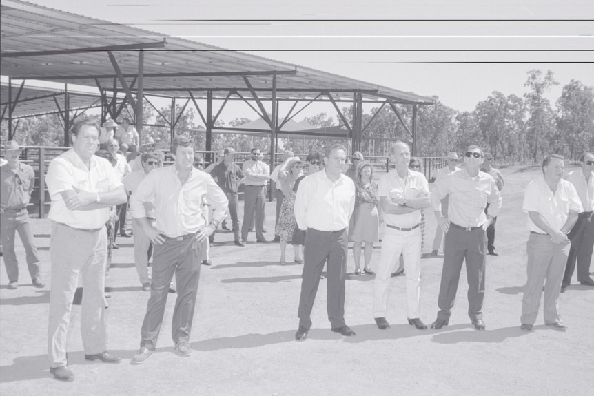 A black and white photo of a group of men stood in the dirt, in front of a large metal structure, the export yard.