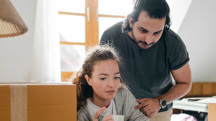 A man and a woman look at a a screen while standing near cardboard boxes.