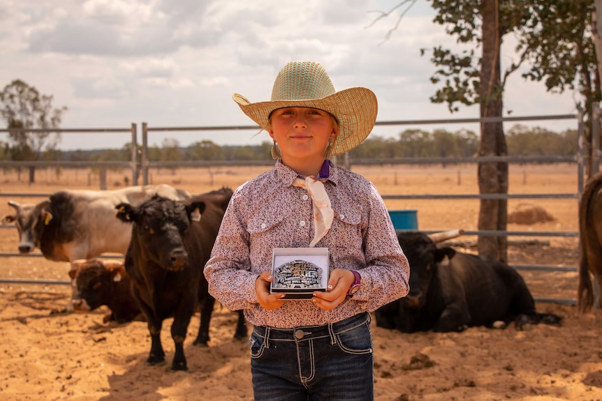 Young girl in big hat stands holding a belt buckle in a box as cattle stand behind her