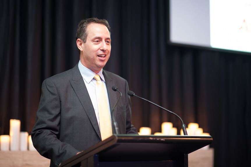 A man in a black suit stands at a podium addressing an audience