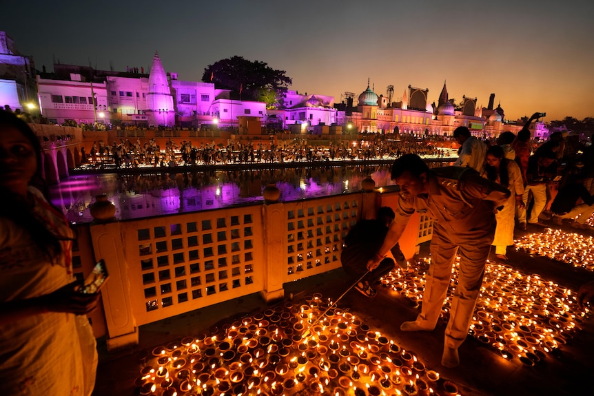 People lean down to light lamps on a balcony over a river. More lanterns and illuminated buildings are on the other side.