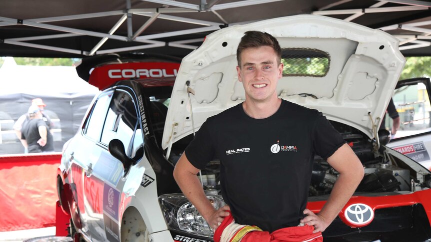 A young man in a black t-shirt standing in front of a rally car with its bonnet up.