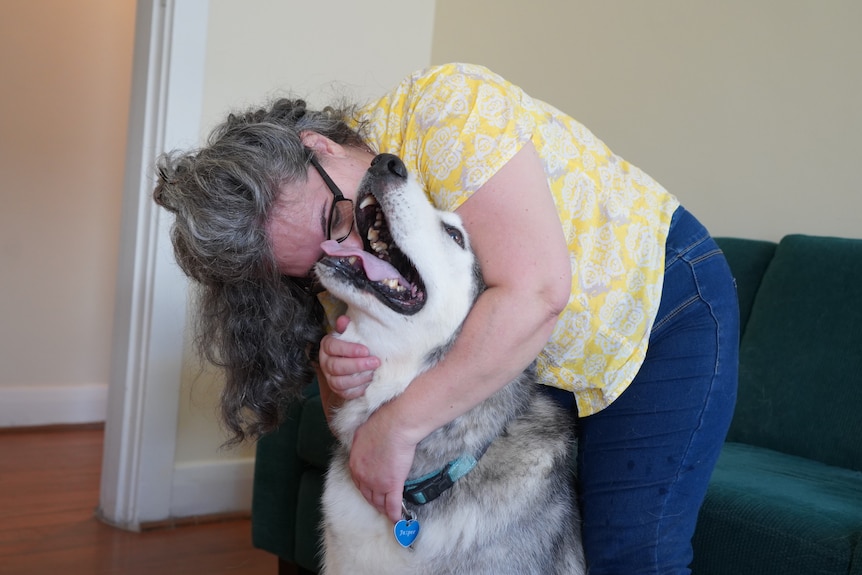 A woman in a yellow shirt with long grey, curly hair cuddles her husky