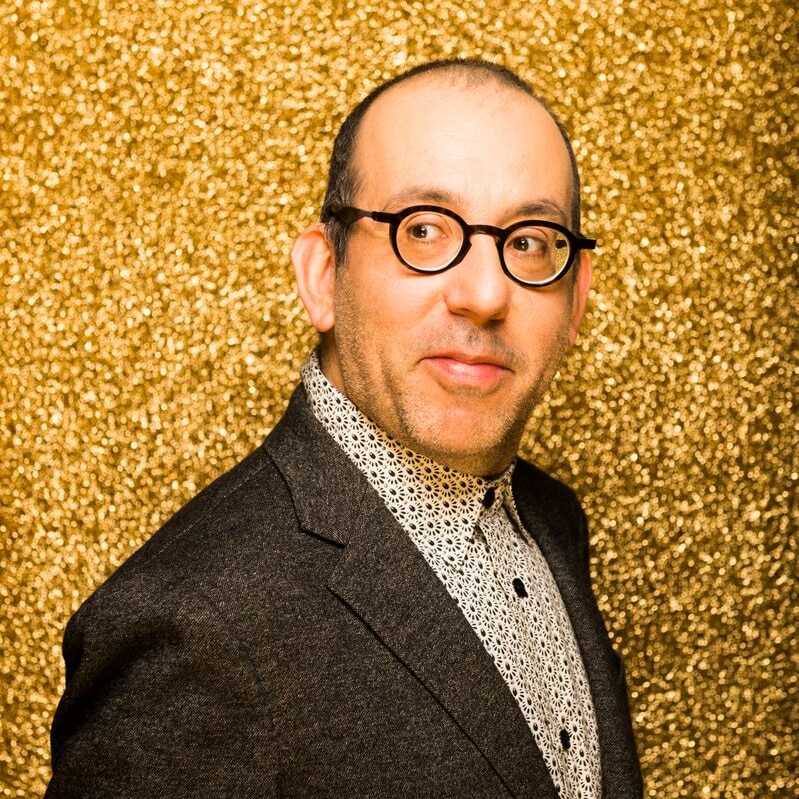 Barrie Kosky wears round glasses, a paisley print shirt and a brown blazer against a gold glittery backdrop.